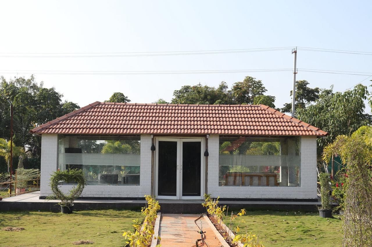Glass-walled dining pavilion with a tiled roof, facing the lawn at Mytri Farmhouse near Kondapochamma Sagar, Siddipet