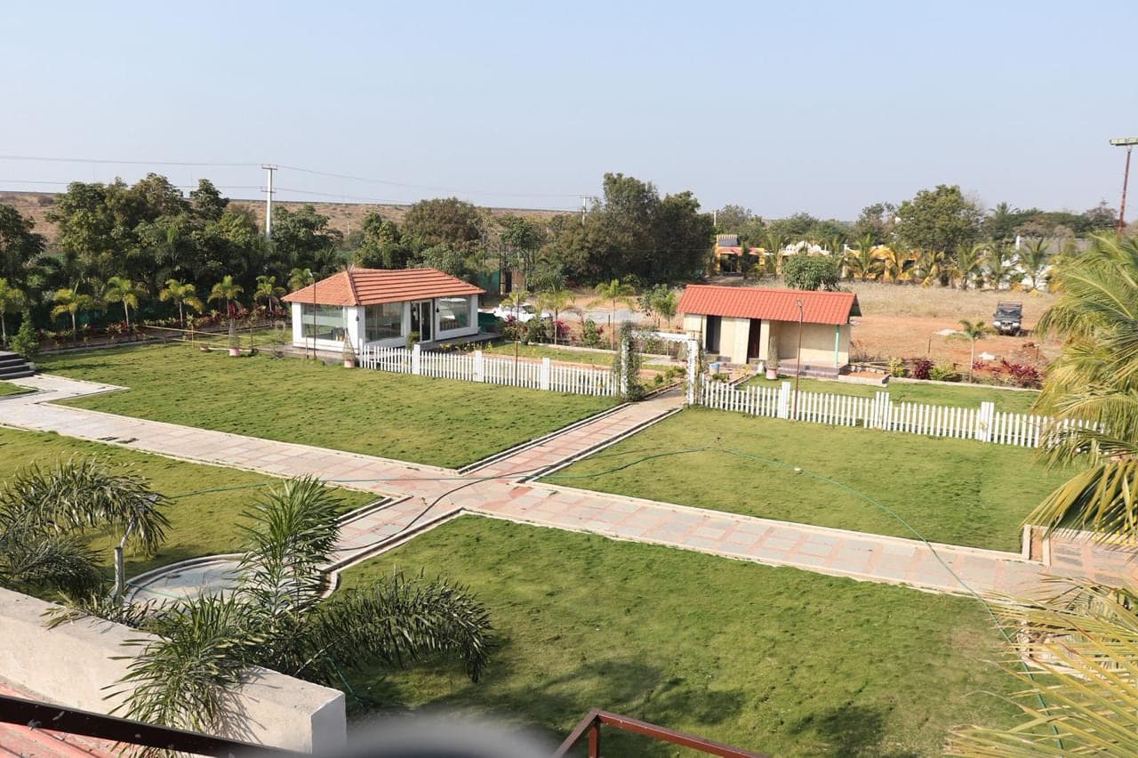 Open lawns, paved paths and the dining pavilion seen from above at Mytri Farmhouse near Kondapochamma Sagar, Siddipet