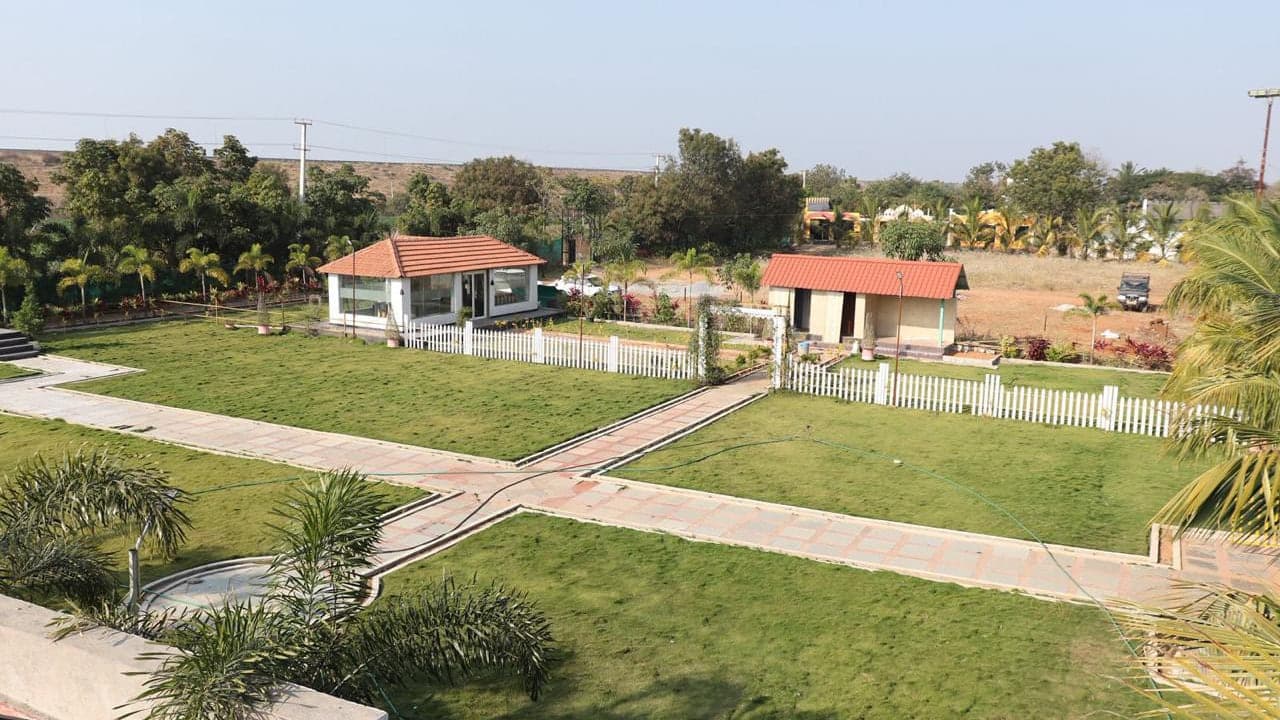 Open lawns divided by paved paths, with the dining pavilion beyond, at Mytri Farmhouse near Kondapochamma Sagar, Siddipet