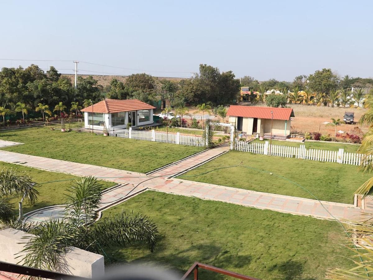 Open lawns divided by paved paths, with the dining pavilion beyond, at Mytri Farmhouse near Kondapochamma Sagar, Siddipet